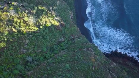 Ascenso-De-Drones-Que-Revela-Un-Acantilado-Verde-Cubierto-De-Vegetación-Tropical-En-Una-Playa-De-Arena-Negra-Volcánica