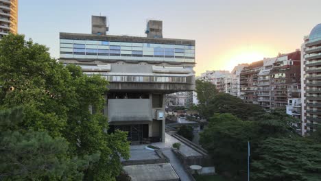 Aerial-parallax-of-brutalist-style-National-Library-surrounded-by-buildings-and-trees-at-sunset,-Buenos-Aires