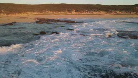 Aerial-backward-flight-over-splashing-ocean-water-crashing-against-rocks-and-ending-at-sandy-beach-during-golden-sunset