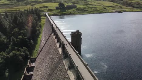 Tourists-crossing-scar-house-reservoir-dam-passage-on-sunny-spring-day,-orbital-aerial-view