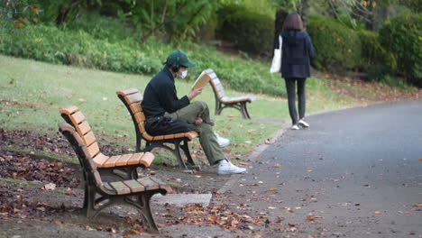 Guy-Wearing-Facemask-Sitting-And-Reading-Book-While-Listening-To-Music-On-Wooden-Bench-In-Park-During-Pandemic