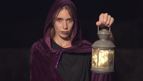 Mysterious-role-play-woman-wearing-a-purple-cape-and-hood-and-holding-a-lantern-at-night