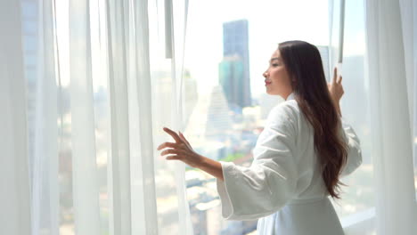 A-pretty-woman-in-a-bathrobe-pulls-back-the-sheer-drapes-in-her-hotel-room-to-reveal-the-view-of-a-modern-city-skyline