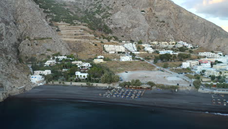 Drohne-Luftaufnahme,-Die-über-Schwarzen-Sandstrand-Mit-Sonnenliegen-Mit-Bergkulisse-Auf-Der-Insel-Santorini,-Griechenland-In-4k-Fliegt