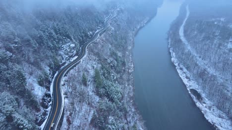Aerial-footage-of-a-snowy,-scenic-byway,-winding-mountain-valley-road-during-a-snowstorm-with-pine-trees,-a-river,-mountain-highway,-rocky-cliffs,-and-forests-during-winter-on-a-cold,-blue-day