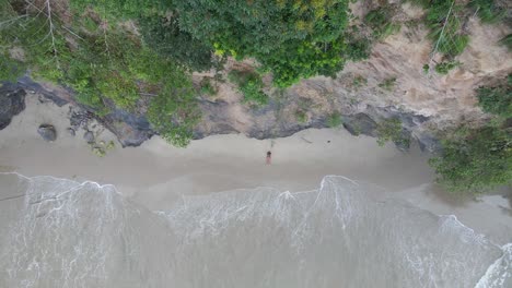Aerial-view-of-a-girl-laying-between-a-cliff-and-waves-of-the-ocean-crashing-at-her-feet