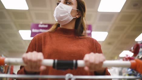 Beautiful-woman-wearing-protective-disposable-medical-mask-and-fashionable-clothes-uses-shopping-cart-while-shopping-in-supermarket