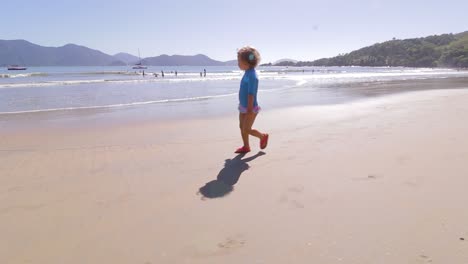 camera-orbiting-around-young-cute-girl-running-freely-on-the-beach-with-mountains-background-and-calm-sea-and-clear-sand-contemplating-the-environment