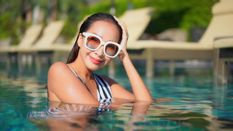 Delighted-lady-half-submerged-in-the-swimming-pool,-wearing-sun-glasses-and-swimwear-smiling-and-looking-at-the-camera
