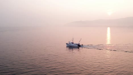Bright-hazy-sunrise-with-fishing-boat-heading-out-to-open-sea,-aerial