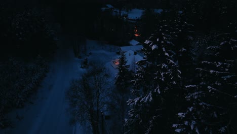 Aerial-view-around-snowy-trees,-revealing-a-illuminated-house,-dark,-winter-evening