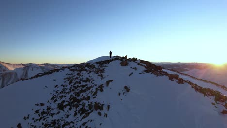 Aerial-View-of-Lonely-Mountaineer-on-Top-of-Rocky-Peak-at-Sunrise,-Snow-and-Rising-Sun-Above-Mountains,-Orbiting-Drone-Shot
