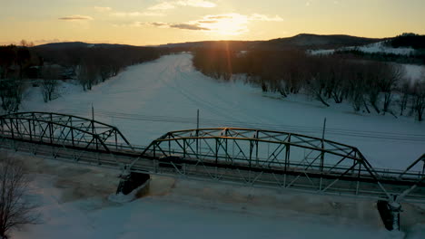 Aerial-view-of-a-car-crossing-a-bridge-over-a-snow-covered-frozen-river-as-the-sun-sets-in-the-winter-sky