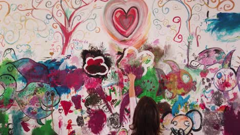 camera-zoom-out-baby-girl-in-pink-dancing-dress-hand-painting-purple-violet-on-colorful-wall-full-of-creative-drawings