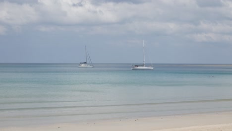 Der-Strand-Mit-Ein-Paar-Yachten-Im-Meer,-Weißer-Sand-Und-Wellen-Vom-Ruhigen-Meer-Bei-Sonnenschein-Tagsüber-In-Phuket