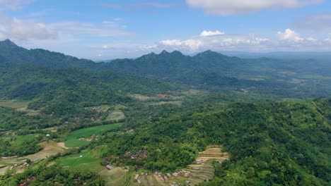 Drone-flying-over-verdant-valley-with-Menoreh-hills-in-background,-Muntilan-in-Indonesia