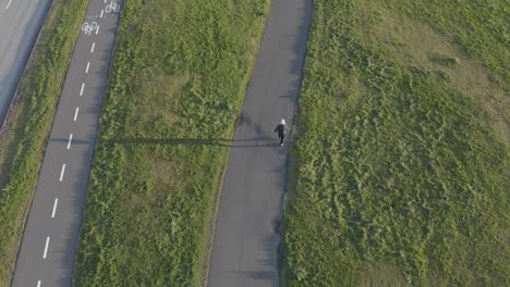 Man-walking-on-sidewalk-surrounded-by-grass,-long-shadow-from-sunset,-aerial