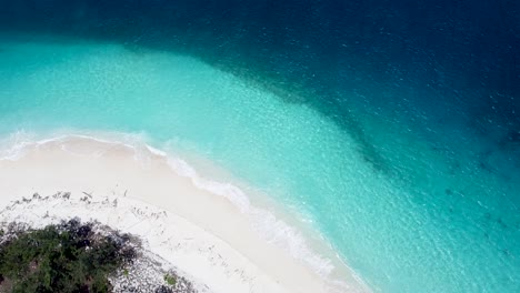 Looking-down-on-a-stunning-remote-tropical-island-with-natural-earth-tones,-white-sandy-beach,-shallow-crystal-clear-water-and-deep-blue-ocean