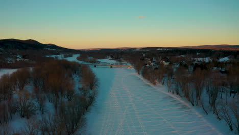 Malerische-Luftaufnahme,-Die-Bei-Sonnenuntergang-Entlang-Eines-Zugefrorenen-Schneebedeckten-Flusses-Fliegt