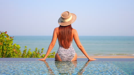 An-attractive-woman-sits-with-her-back-to-the-camera-on-the-edge-of-an-infinity-edge-pool,-looking-out-on-the-ocean-and-the-incoming-waves