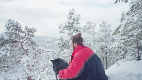 Man-With-His-Alaskan-Malamute-Dog-Sitting-On-Cold-Winter-Day-In-Norway---medium-shot