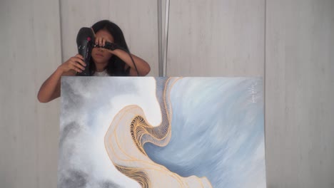 Female-Artist-Drying-Top-Side-Of-A-Paint-Canvas-With-A-Hair-Dryer-In-The-Studio