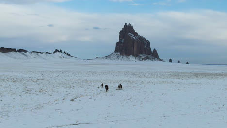 Wild-horses-grazing-in-snow-in-front-of-Shiprock-mountain-formation,-New-Mexico,-USA