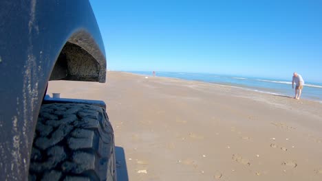 Right-front-tire-of-vehicle-and-people-walking-in-the-surf-while-driving-on-a-beach-on-a-sunny-day-on-South-Padre-Island-Texas--Point-of-view,-POV