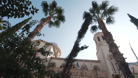 Tall-exotic-Palm-Trees-parallel-to-lateral-aisle-Agia-Napa-Cathedral-Towers-in-Limassol,-Cyprus---Wide-low-angle-slow-rotation-shot