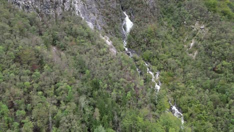 Waterfall-cascading-through-trees-Lake-Bohinj-Slovenia-pull-back-reveal-drone-aerial-view