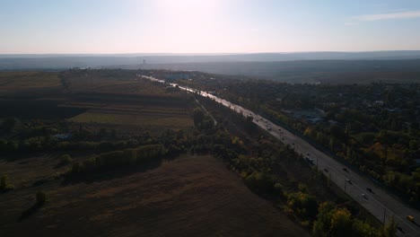 Panoramic-aerial-drone-flight-into-the-sun-along-the-Hincesti-Hwy-M3-highway-south-bound-with-heavy-traffic-in-the-capital-Chisinau,-Republic-of-Moldova,-2022---autumn-with-colorful-fields-and-trees