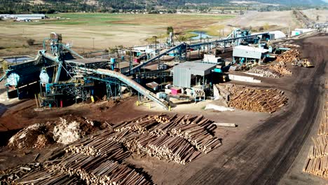 dolly-forward-zoom-drone-shot-of-a-blue-Sawmill-Processing-Plant-with-log-piles-in-the-background-in-a-desert-environment-with-heavy-equipment-moving