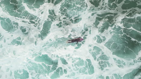 Aerial-top-down-shot-of-a-surfer-paddling-through-thick-white-foam-and-over-a-swell-of-an-incoming-wave-as-he-heads-back-out-to-the-backline,-Suances,-Spain