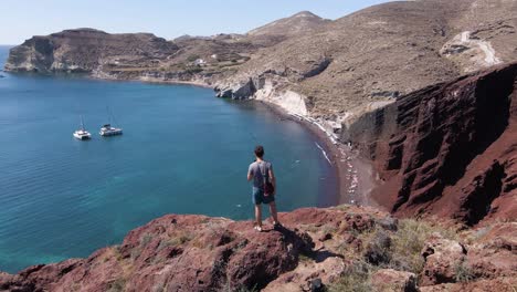Aerial-shot-over-male-person-standing-at-cliff-edge-over-Red-Beach-in-Santorini,-Greece