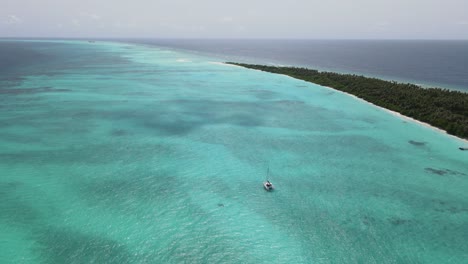 Drone-shot-of-turquoise-indian-ocean-with-small-yacht-sailing-on-calm-waters-near-tropical-maldivian-island