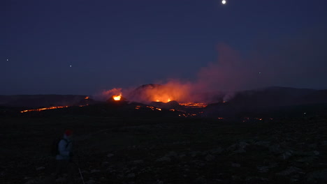 Weite-Aufnahme-Eines-Unglaublichen-Vulkanausbruchs-Im-Hintergrund-Und-Eines-Wandernden-Touristen-Mit-Taschenlampen-In-Der-Nacht-Im-Vordergrund