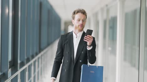 Handsome-adult-man-walking-down-the-hall-with-shopping-bags-and-a-smartphone