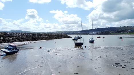 Aerial-view-moored-boats-on-Welsh-low-tide-seaside-breakwater-harbour-coastline-pull-back-descending-low