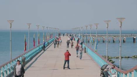 Menschen,-Die-Auf-Der-Palanga-Brücke-Spazieren-Gehen,-Während-Fahnen-Im-Wind-Wehen-Und-Die-Ostsee-Ruhig-Unter-Dem-Pier-Plätschert