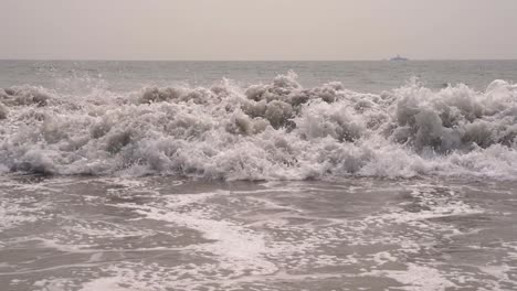 Close-up-of-Pacific-ocean-waves-crashing-down-onto-the-sandy-beach-in-slow-motion-in-San-Diego,-California-with-a-large-ship-in-the-background