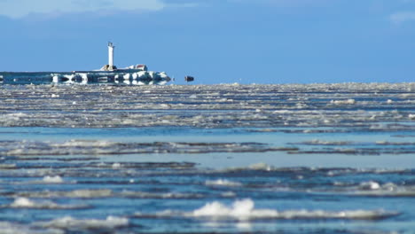 Melted-ice-flows-down-sunny-river-in-winter-with-lighthouse,-long-shot