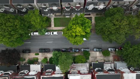 Tracking-shot-of-white-car-driving-down-quaint-suburban-street-lined-with-townhomes,-parked-cars,-and-trees