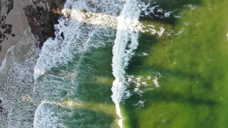 A-lime-green-ocean-mixed-with-hints-of-dark-blue-pushes-small-waves-into-the-dark-rocks-sat-between-the-pale,-coarse-sand-whilst-shadows-cast-from-apartment-buildings-block-the-sunsets-afternoon-glow