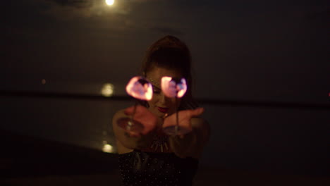 Woman-Twirling-Fire-Palm-Torches-Against-Full-Moon-lit-Lake,-Night-Exterior-Medium-Shot-Slowmo