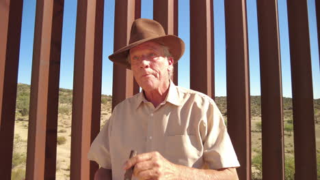 American-Male-Guy-with-fedora-hat-lights-cigar-in-front-of-US-Mexico-border-fence