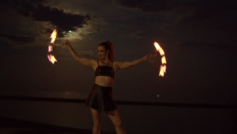 Woman-Twirling-With-Spinning-Fire-Fans-On-Lakeshore-Beach,-Night-Exterior-Slowmo