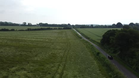 drone-car-follow-shot-over-a-wet-field,-the-car-to-the-right-of-the-shot-is-speeding-down-a-narrow-country-lane-between-two-fields-on-an-overcast-day-in-england