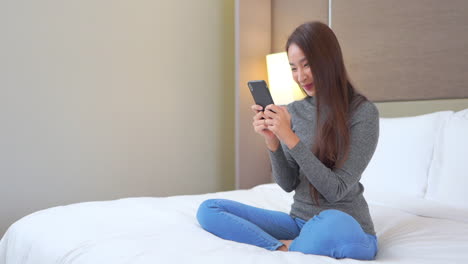 A-pretty-young-woman-sitting-on-a-bed-reacts-with-a-laugh-to-something-on-her-smartphone