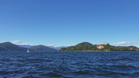 Pequeño-Velero-Navegando-En-El-Lago-Maggiore-Aguas-Agitadas-Con-Montañas-Y-Castillo-De-Angrya-En-El-Fondo,-Italia