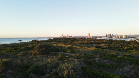 Low-fast-drone-view-travelling-over-a-coastal-nature-parklands-with-a-urban-city-skyline-rising-above-in-the-foreground